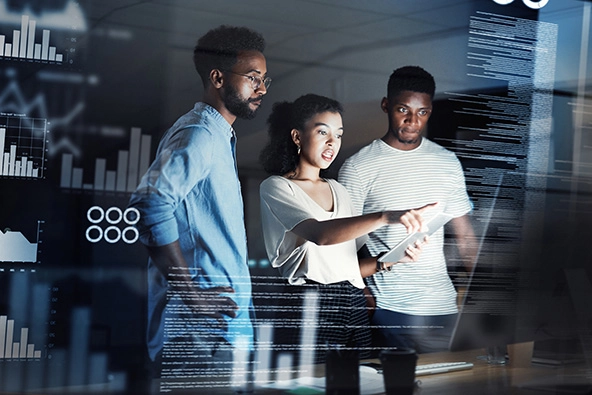 Three young programmers having a discussion together while standing and working next to a computer inside of their office at night