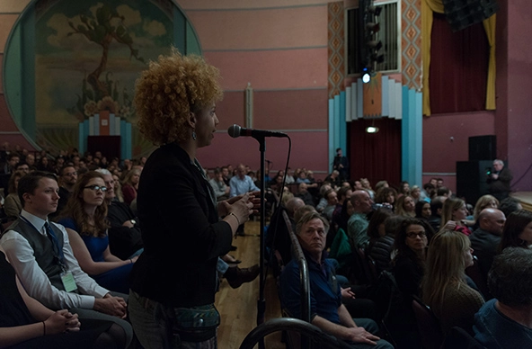 Black woman standing and speaking into a microphone in a crowd of people