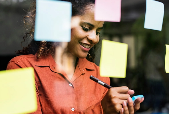 Woman writing on a sticky note, standing in front of wall of sticky notes