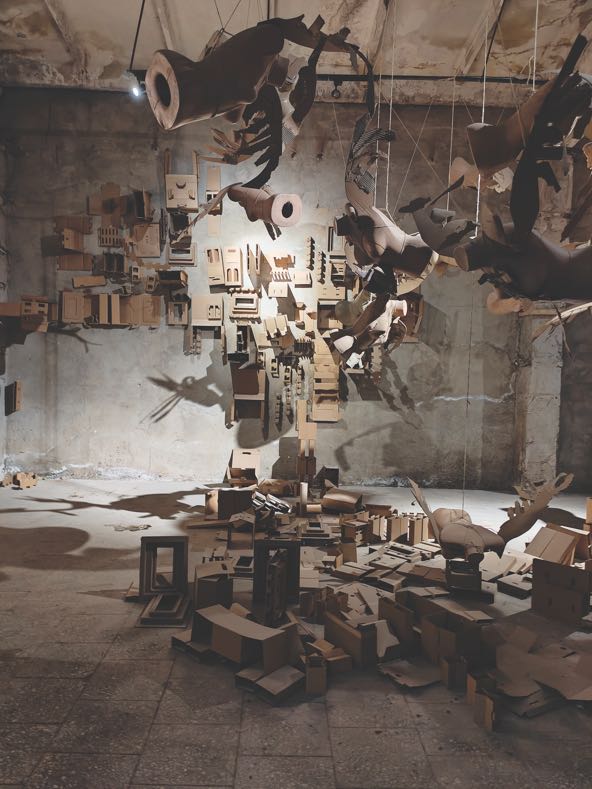 sculpture made of many tan to brown geometrical shapes and mannequin parts that is  suspended from the ceiling or resting on the floor in a museum