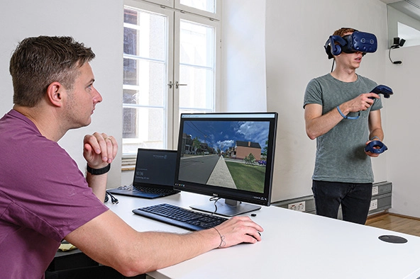 Man at a computer watching someone wearing a virtual reality headset
